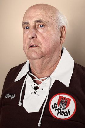 Portrait von Dieter Rittmeyer, dem Gründer des FC St. Pauli-Archivs. Das Foto zeigt einen alten Mann in einem altmodischen braun-weißen FC St. Pauli-Trikot.