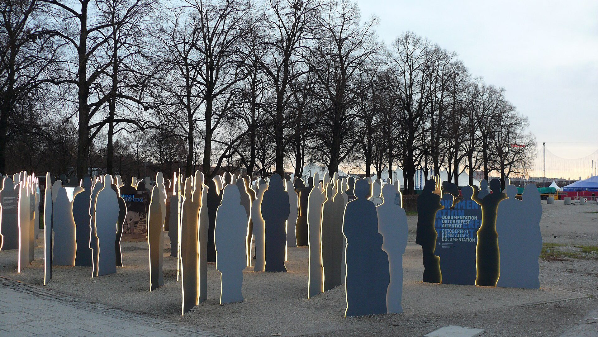 Zu sehen ist ein Foto: Am Nordrand der Theresienwiese erinnert dieses Denkmal an das Attentat auf das Oktoberfest am 26. September 1980: Östlich der Parkplatz-Einfahrt: eine Installation elektrisch beleuchteter Pappkameraden (2020), offiziell bezeichnet als „Dokumentation Oktoberfest-Attentat“, aber die künstlerische Gestaltung weist die Anlage – trotz des Namens und des „unedlen“ Materials – natürlich dennoch als Denkmal aus.