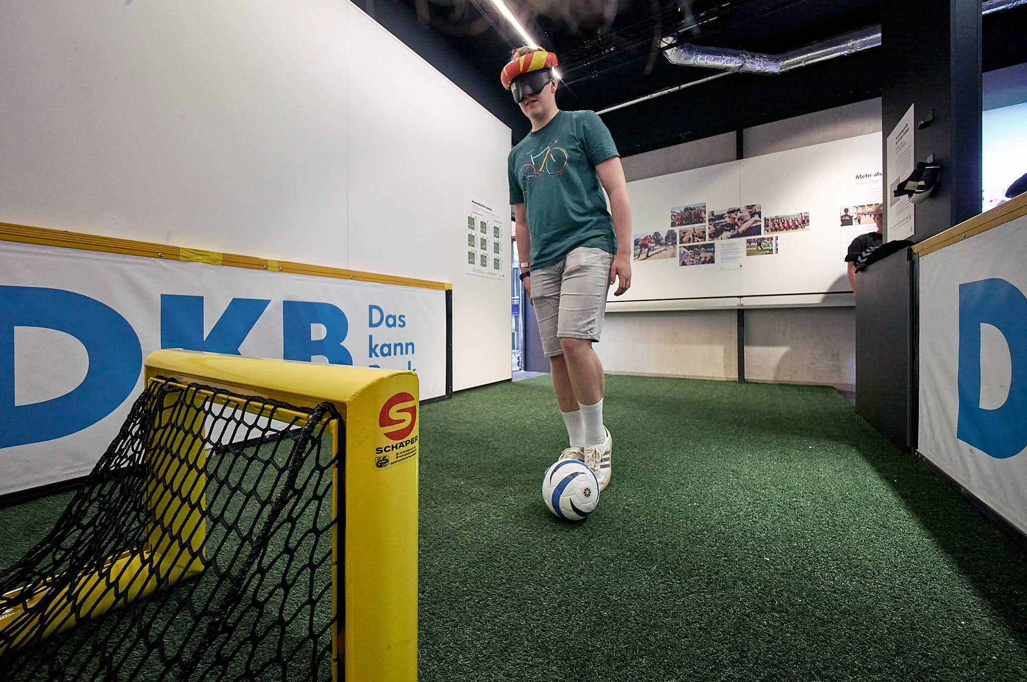 Eine Person steht in einer interaktiven Mitmach-Station der Ausstellung, trägt eine Verdunklungsbrille und agiert mit dem Ball am Fuß vor einem Tor.