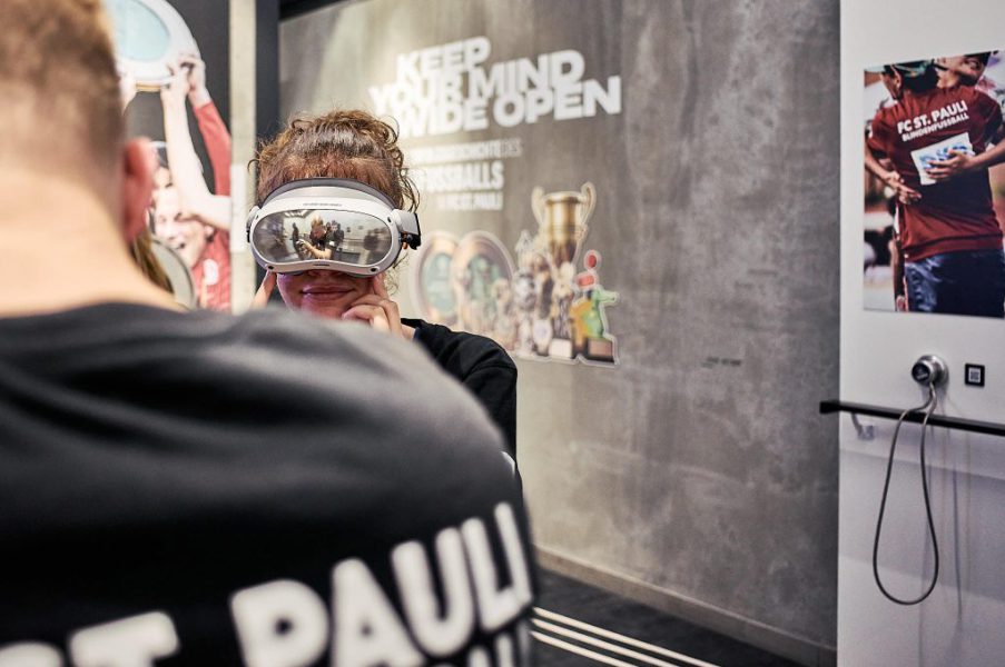 Hier ist eine Blindenfußballspielerin zu sehen, die in der Ausstellung steht und eine VR-Brille trägt.